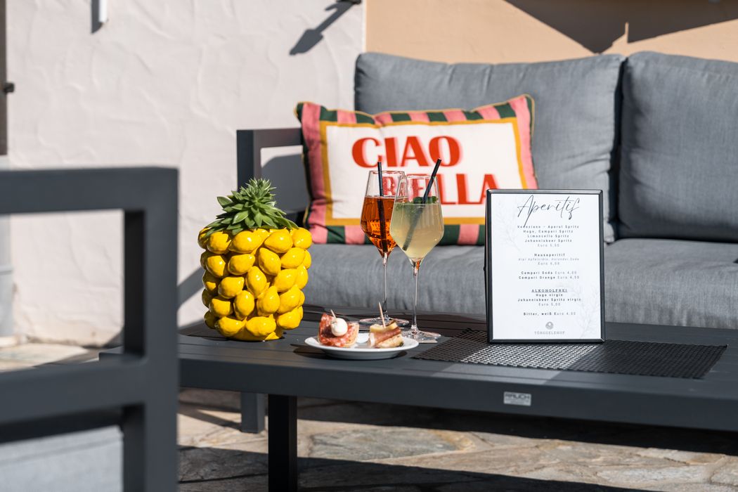 Sommerliche Terrasse mit grauem Sofa, Kissen mit Aufschrift 'Ciao Bella', gelber Dekopflanze, Getränken und Aperitifkarte auf schwarzem Tisch. Sommerliche Terrasse mit grauem Sofa, Kissen mit Aufschrift 'Ciao Bella', gelber Dekopflanze, Getränken und Aperitifkarte auf schwarzem Tisch.