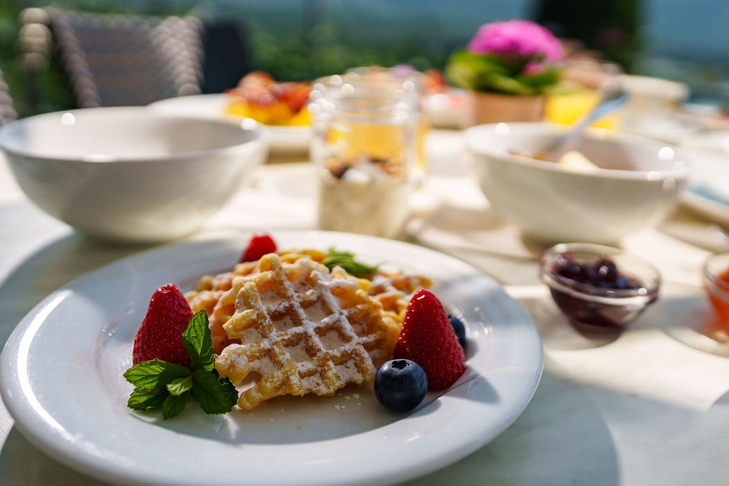 Waffeln mit Puderzucker, Erdbeeren, Blaubeeren und Minzblättern auf einem weißen Teller, serviert auf einem Tisch mit weiteren Frühstücksgerichten im Hintergrund. Waffeln mit Puderzucker, Erdbeeren, Blaubeeren und Minzblättern auf einem weißen Teller, serviert auf einem Tisch mit weiteren Frühstücksgerichten im Hintergrund.