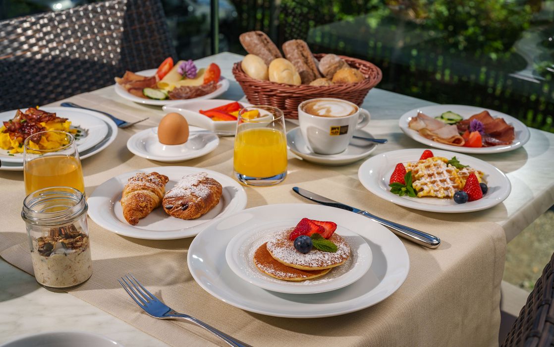 Tisch mit reichhaltigem Frühstück, darunter Pancakes mit Früchten, Croissants, Waffeln, Orangensaft, Kaffee, Ei und Brotkorb im Freien Tisch mit reichhaltigem Frühstück, darunter Pancakes mit Früchten, Croissants, Waffeln, Orangensaft, Kaffee, Ei und Brotkorb im Freien