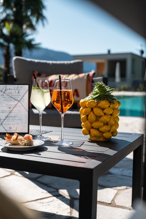 Sommerliche Terrasse mit zwei gefüllten Weingläsern, einer Ananas-Dekoration, kleinen Häppchen auf einem Teller und einem Aperitif-Menü, im Hintergrund Pool und Palmen. Sommerliche Terrasse mit zwei gefüllten Weingläsern, einer Ananas-Dekoration, kleinen Häppchen auf einem Teller und einem Aperitif-Menü, im Hintergrund Pool und Palmen.
