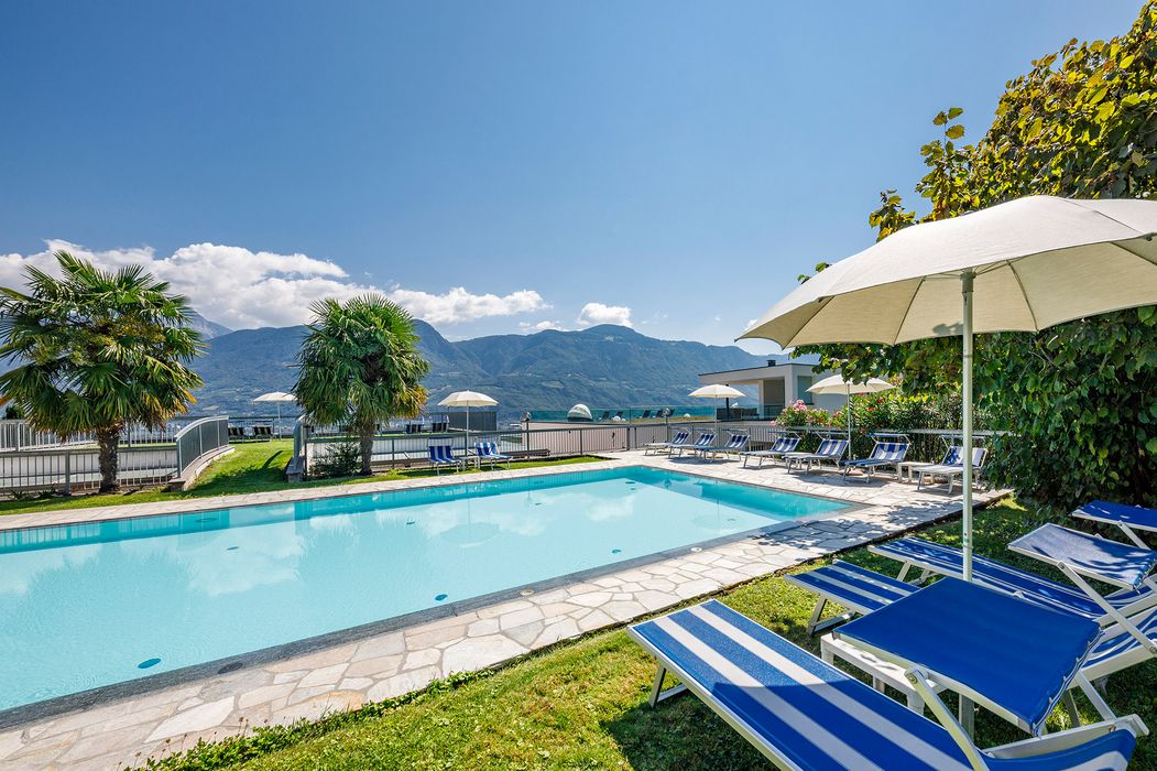 Freier Poolbereich mit blauen Liegestühlen und weißen Sonnenschirmen vor Bergkulisse und klarem Himmel.