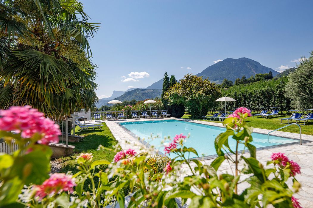 Schwimmbad in einer Gartenanlage mit Liegestühlen, Sonnenschirmen, bunten Blumen und Bergen im Hintergrund bei klarem Himmel