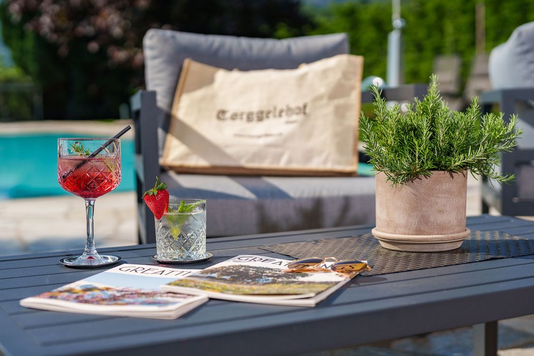 Tavolo da esterno con due drink, riviste, occhiali da sole e pianta in vaso, con poltrona e piscina sullo sfondo