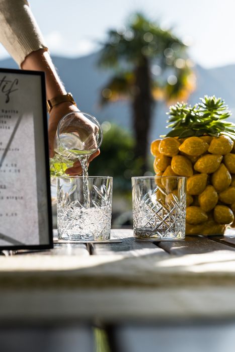 Person gießt Wasser in ein Kristallglas auf einem Holztisch mit einer Dekoration aus gelben Zitronen im Hintergrund. Person gießt Wasser in ein Kristallglas auf einem Holztisch mit einer Dekoration aus gelben Zitronen im Hintergrund.