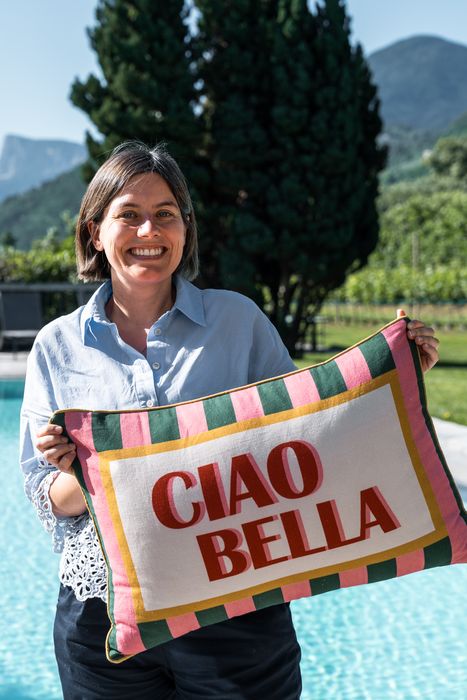 Frau lächelt draußen am Pool und hält ein buntes Kissen mit der Aufschrift 'CIAO BELLA'