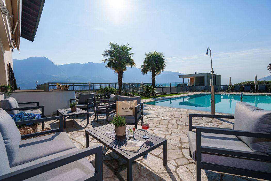 Area piscina con sedie e tavoli grigi, due palme e montagne sullo sfondo sotto un cielo sereno.