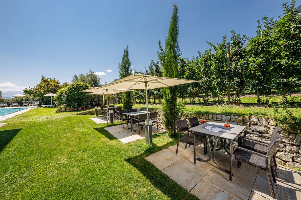 Garten mit Tischen, Stühlen und Sonnenschirmen neben einer Obstplantage unter blauem Himmel