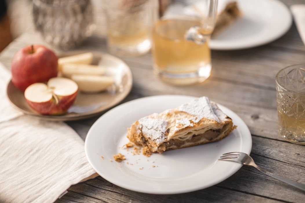 Ein Stück Apfelstrudel auf weißem Teller mit einer Gabel, im Hintergrund ein Teller mit frischen Apfelstücken und Getränke auf Holztisch. Ein Stück Apfelstrudel auf weißem Teller mit einer Gabel, im Hintergrund ein Teller mit frischen Apfelstücken und Getränke auf Holztisch.