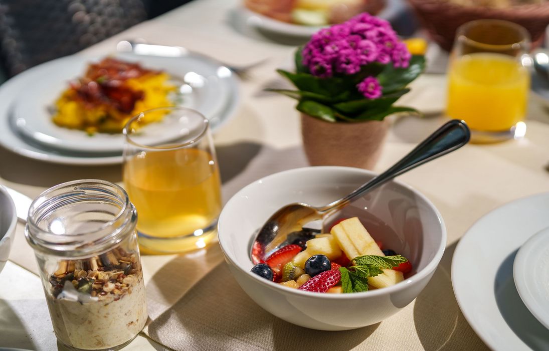 Tisch mit Frühstück, darunter eine Schüssel mit frischem Obst und einer Minzblattgarnierung, ein Glas Saft, ein Glas mit Müsli und ein Blumenarrangement. Tisch mit Frühstück, darunter eine Schüssel mit frischem Obst und einer Minzblattgarnierung, ein Glas Saft, ein Glas mit Müsli und ein Blumenarrangement.