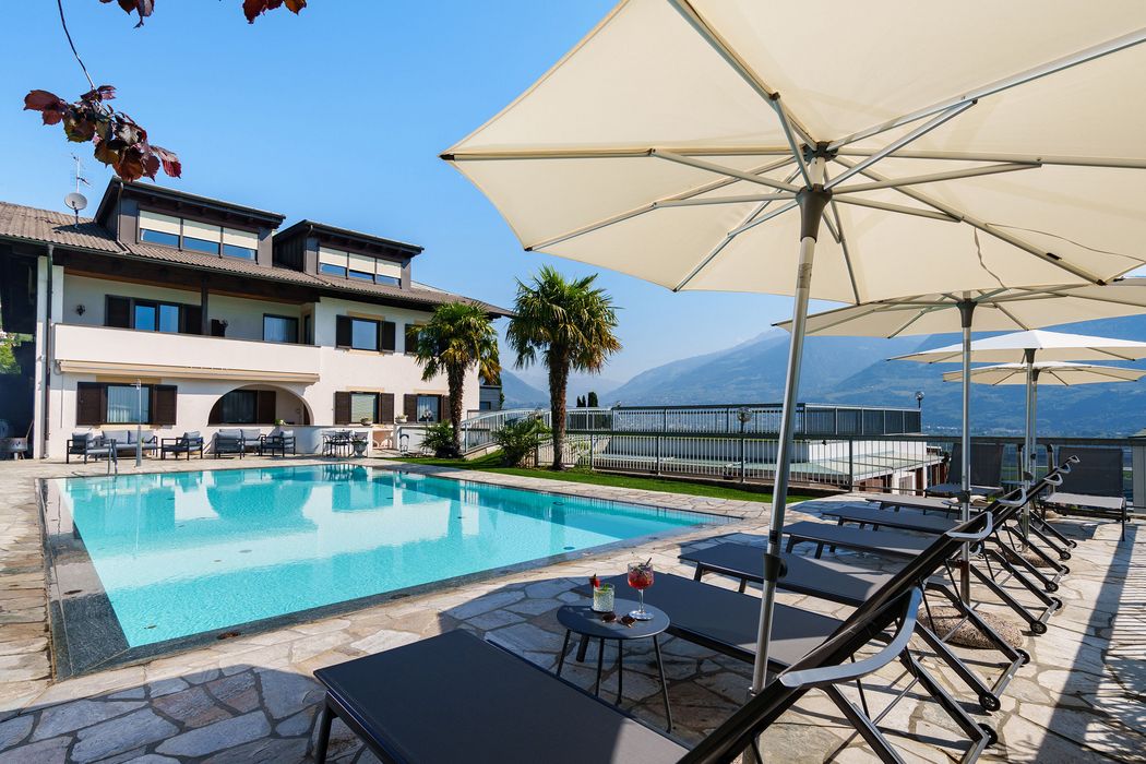 Piscina all'aperto con lettini e ombrelloni in un cortile soleggiato con casa e montagne sullo sfondo