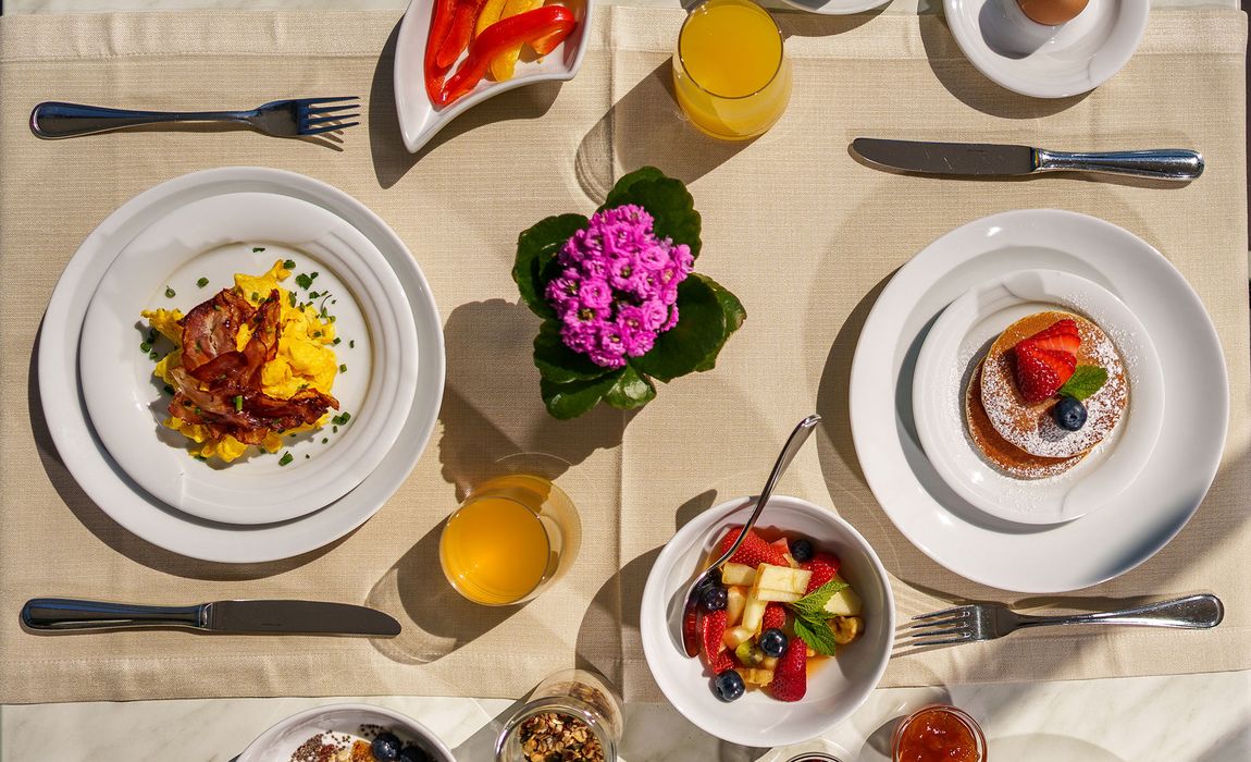 Frühstückstisch mit Rührei und Speck, Obstsalat, Pancakes mit Beeren, Orangensaft und einer Blume in der Mitte Frühstückstisch mit Rührei und Speck, Obstsalat, Pancakes mit Beeren, Orangensaft und einer Blume in der Mitte