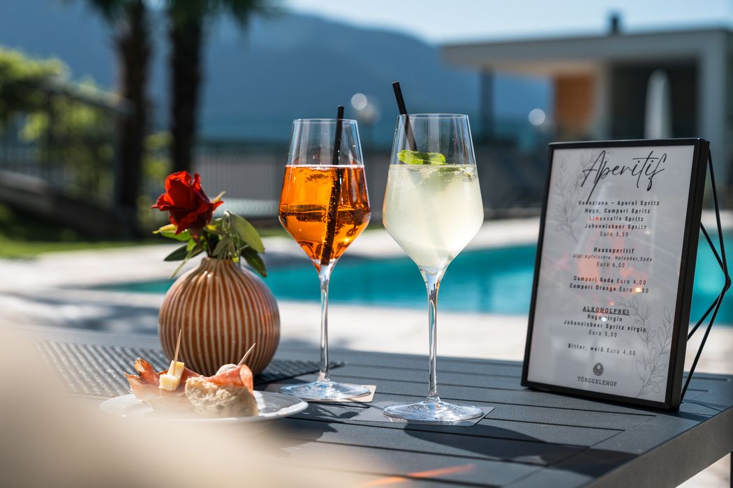 Zwei Aperitif-Gläser mit Strohhalmen, rote Rose in Vase, Smallplate mit Snacks und Aperitif-Menü am Tisch neben Pool im Freien Zwei Aperitif-Gläser mit Strohhalmen, rote Rose in Vase, Smallplate mit Snacks und Aperitif-Menü am Tisch neben Pool im Freien